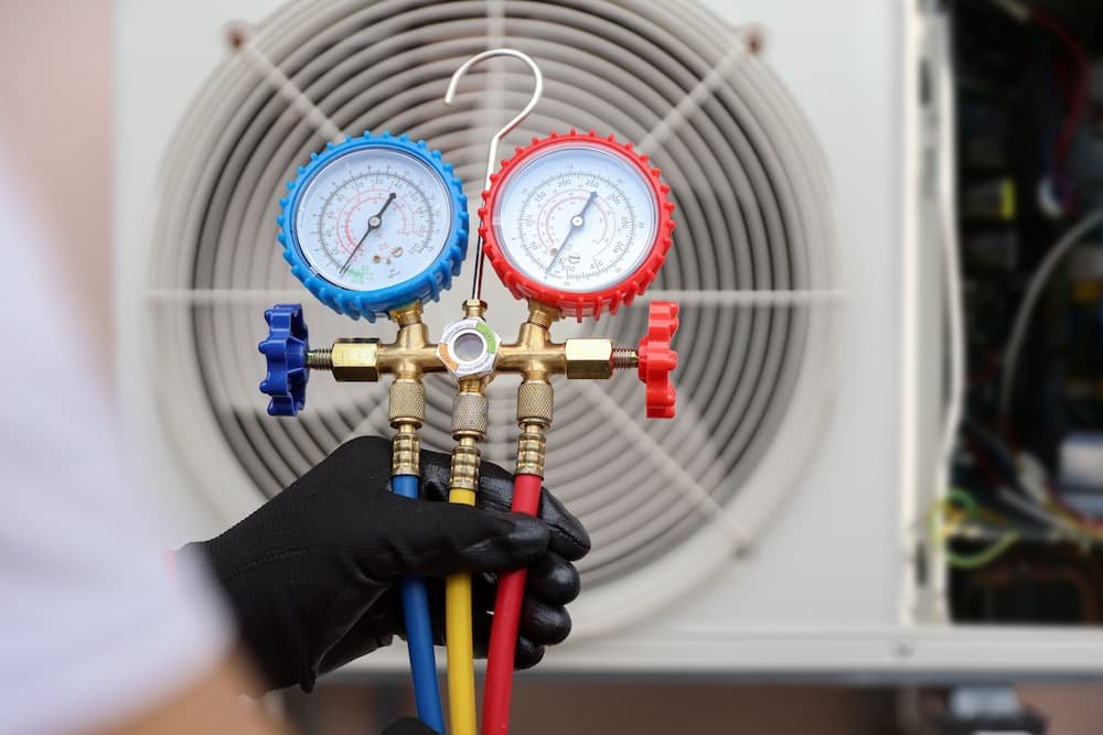 HVAC technician holding a manifold gauge set with blue and red pressure dials in front of an outdoor air conditioning condenser unit.